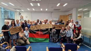 Image is a Group Photo from the 8th ESTHER Ireland Partnerships Forum 2023. Showing all attendees and staff, with those in the middle holding up wooden sculptures depicting the care pathway of pneumonia in Malawi, as well as the flag of Malawi.